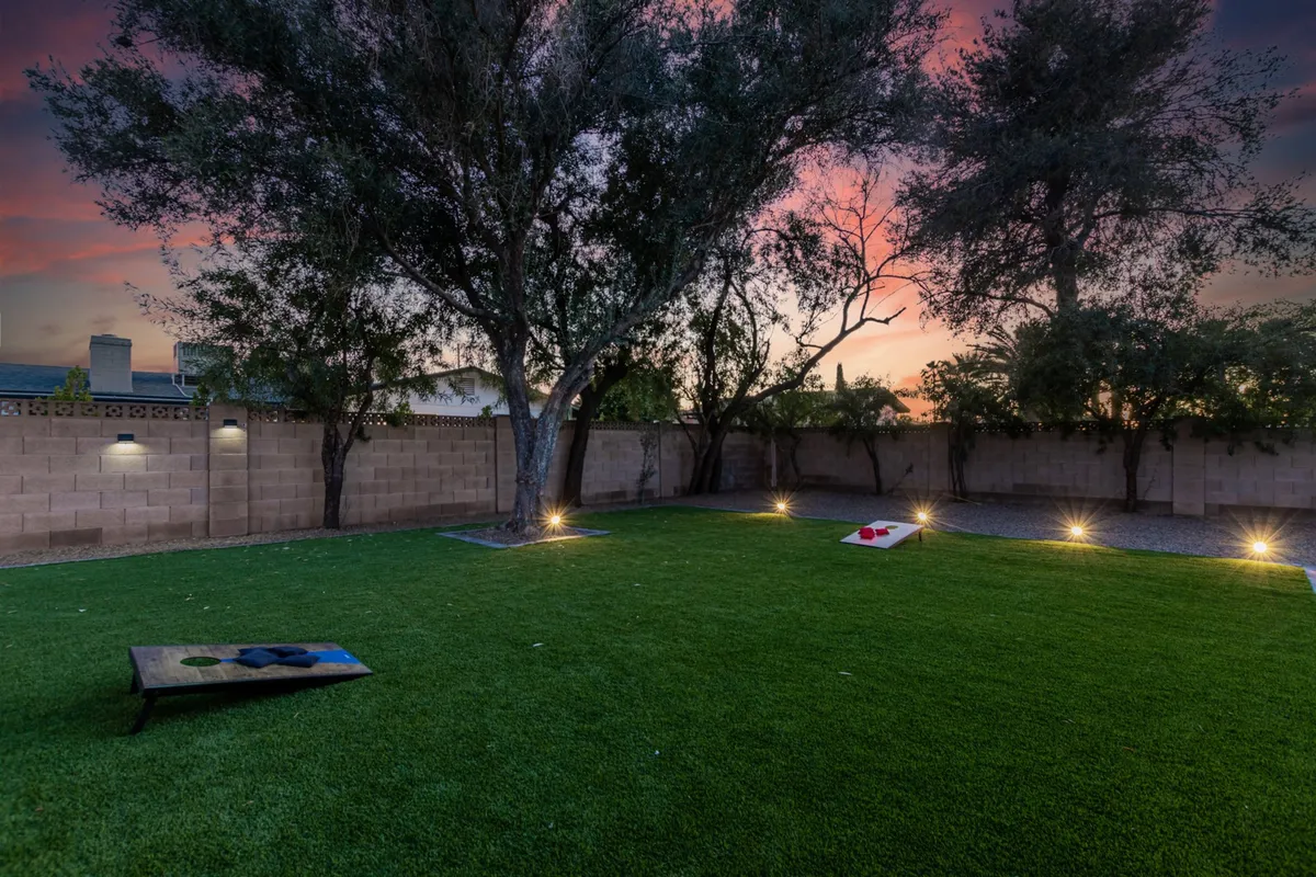 A well-lit grassy space featuring cornhole boards, mature trees, and warm evening lighting, perfect for friendly games or relaxing under the stars. The scene emphasizes the playful and pet-friendly nature of the yard.