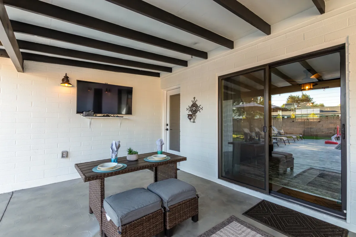 A cozy outdoor dining setup featuring a wicker table, cushioned stools, a wall-mounted TV, and sliding glass doors leading to the interior. The space is illuminated by natural light and arranged for casual gatherings and evening relaxation.