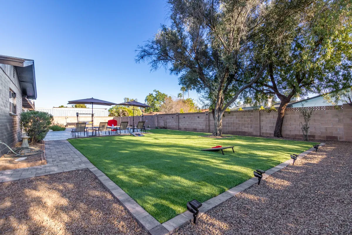 A lush green lawn extends beneath the shade of mature trees, with a casual patio seating area and an outdoor umbrella set up for lounging or socializing. The scene radiates an inviting, relaxed vibe, perfect for enjoying with a pet. The openness of the space encourages outdoor games or simply relaxing with a furry companion under the leafy canopy.