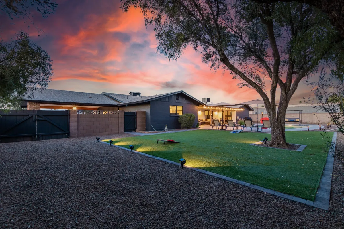 A lush green lawn sits under a large tree, with a cozy outdoor seating area perfectly positioned for sunset views. The vibrant sunset sky creates a dynamic desert backdrop, ideal for gatherings, playtime, and unwinding under open skies.