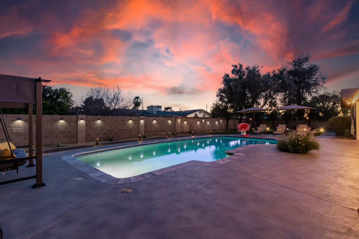 A tranquil swimming pool is framed by a colorful sunset sky, a well-furnished patio area with comfortable chairs, and lush landscaping. The peaceful atmosphere highlights opportunities for both relaxation and inspiring outdoor work sessions.