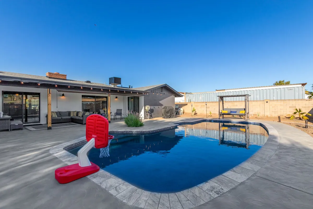 A vibrant outdoor scene featuring a sparkling swimming pool with a basketball hoop, surrounded by an inviting outdoor seating area and a covered patio. The space is filled with natural light and offers a true sense of backyard fun and relaxation, perfect for swimming, sunbathing, and entertaining.