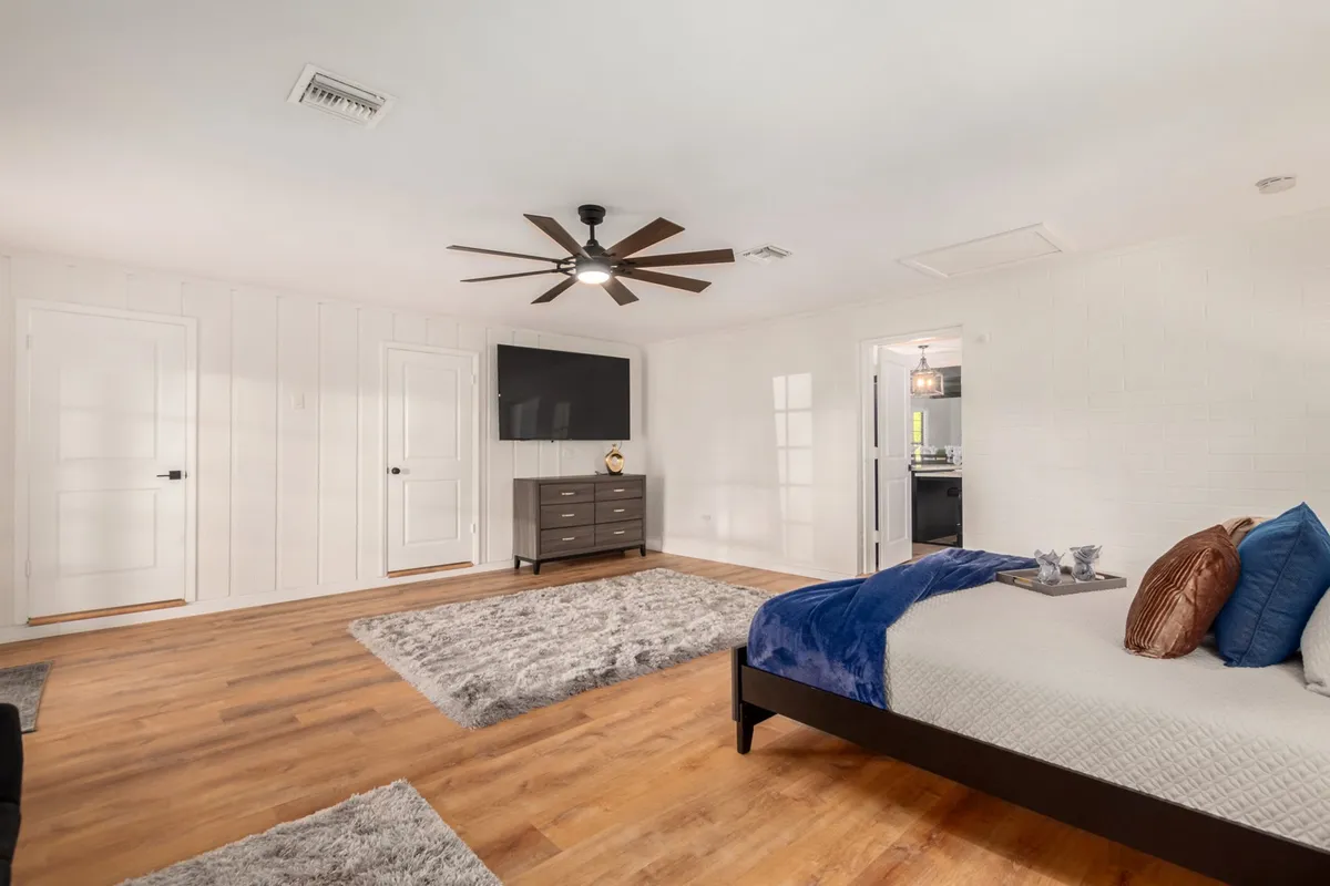 A cozy bedroom featuring a king-sized bed with blue and brown accents, a large wall-mounted TV, dark wood dresser, and a ceiling fan. The room is softly lit and has a modern, serene vibe, showcasing plush bedding and an inviting atmosphere.