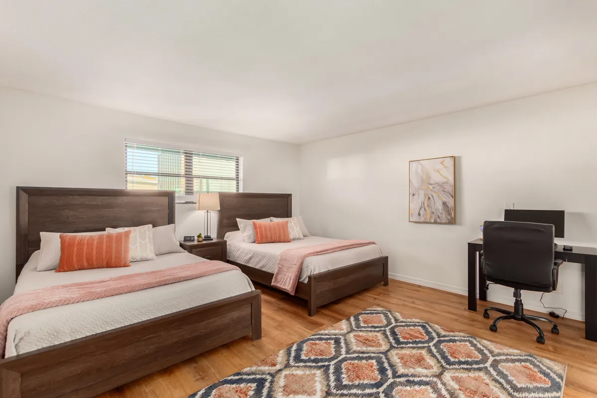 A well-lit bedroom with two beds, a desk and chair, and a decorative art piece. The room is both cozy and functional, suitable for relaxing, studying, or remote work.
