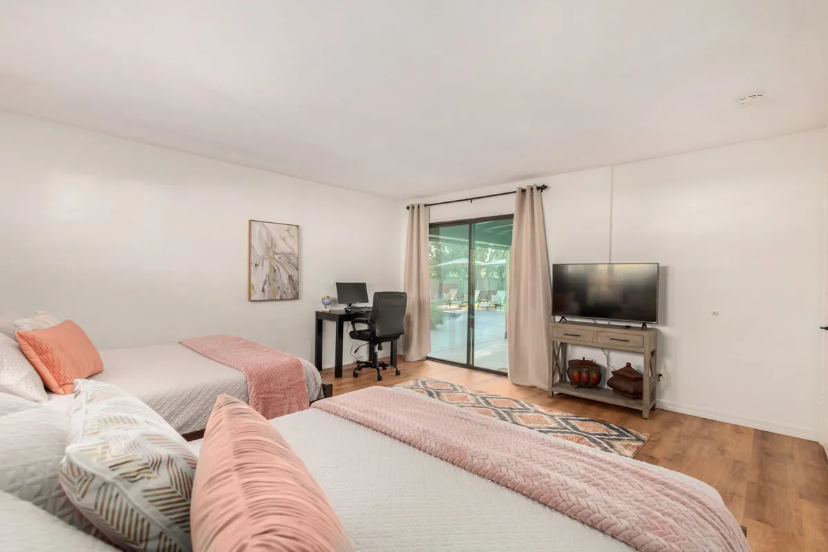 A spacious bedroom with two neatly made beds, a desk and chair for work or study, a television, and a sliding glass door that opens directly to the backyard. The space is filled with natural light, offering a bright and welcoming environment for families or groups.