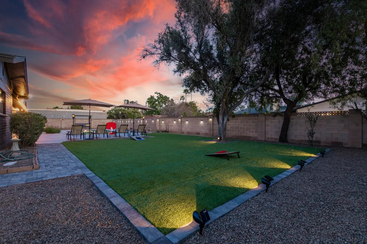 A lush green lawn stretches across the backyard, with a stylish patio seating area and glowing decorative lighting set under a dramatic, colorful sunset sky. The space invites outdoor games like cornhole and gatherings, exuding a relaxed, cheerful ambiance.