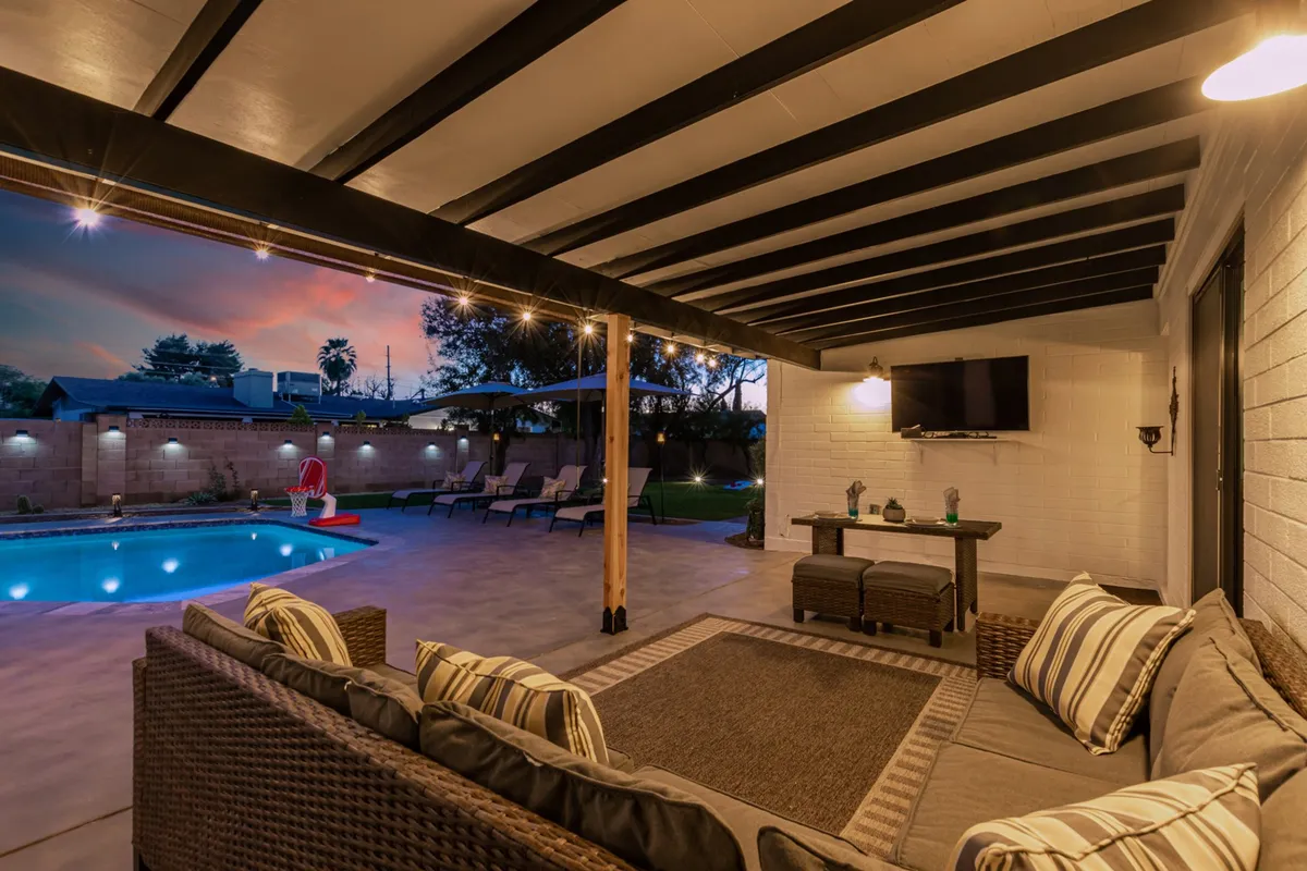 This inviting exterior scene showcases a well-appointed outdoor living area featuring a sparkling swimming pool, comfortable seating, glowing string lights, a mounted outdoor television, and lush landscaping. The space is clearly designed for both lively gatherings and quiet relaxation—ideal for post-game celebrations or decompressing after an exciting day at the ballpark.